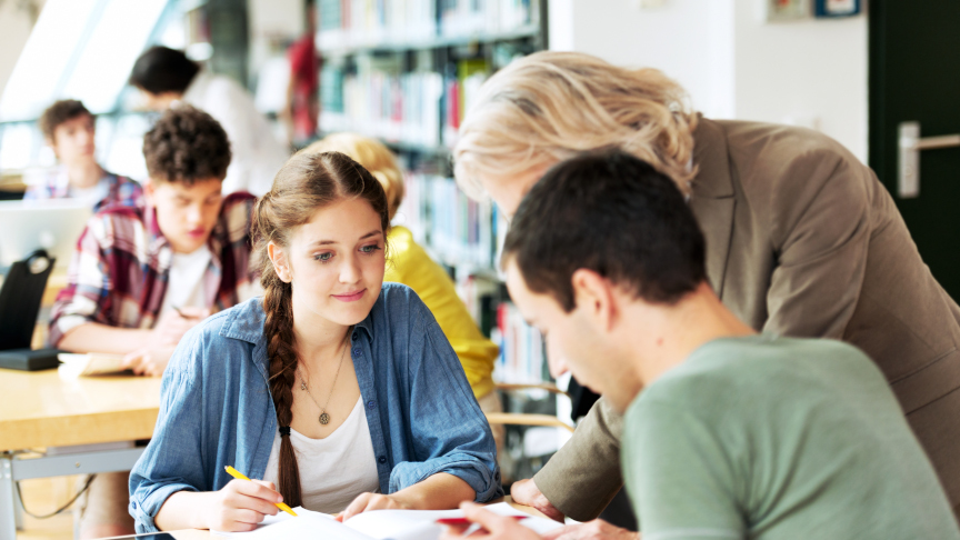 International students sit at tables in the library, a lecturer helps them study.